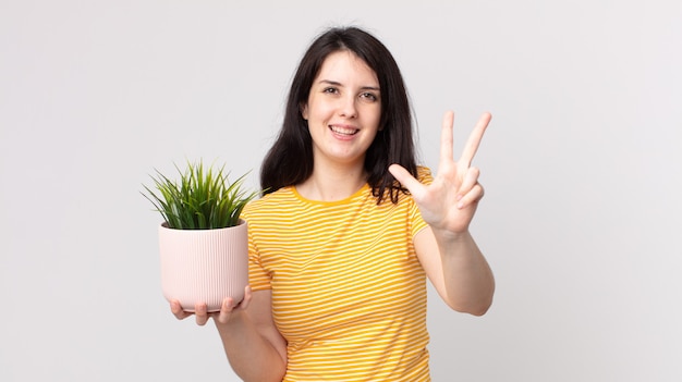 Pretty woman smiling and looking friendly, showing number three and holding a decorative plant
