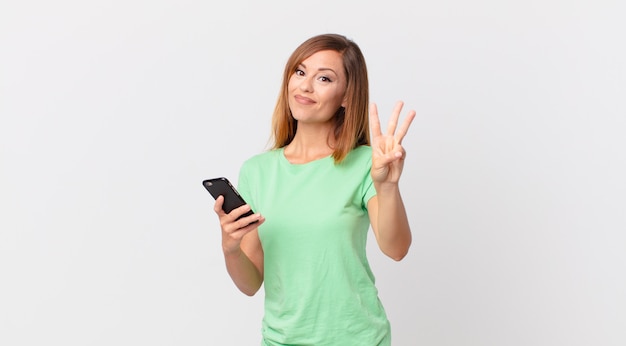 Pretty woman smiling and looking friendly, showing number three and using a smartphone