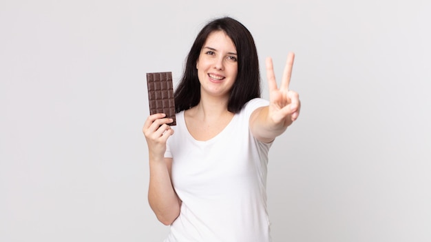 Pretty woman smiling and looking friendly, showing number two and holding a chocolate bar