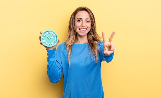 Pretty woman smiling and looking happy gesturing victory or peace holding an alarm clock