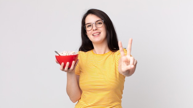 Pretty woman smiling and looking happy, gesturing victory or peace and holding a breakfast bowl