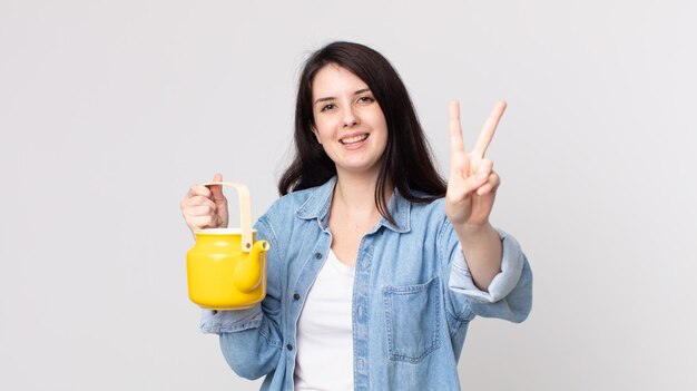 Pretty woman smiling and looking happy, gesturing victory or peace and holding a teapot