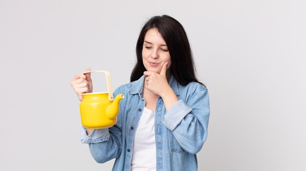 Pretty woman smiling with a happy, confident expression with hand on chin and holding a teapot