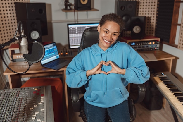 Pretty woman sound engineer gestures showing a heart shape from\
fingers the sound recording or broadcasting studio