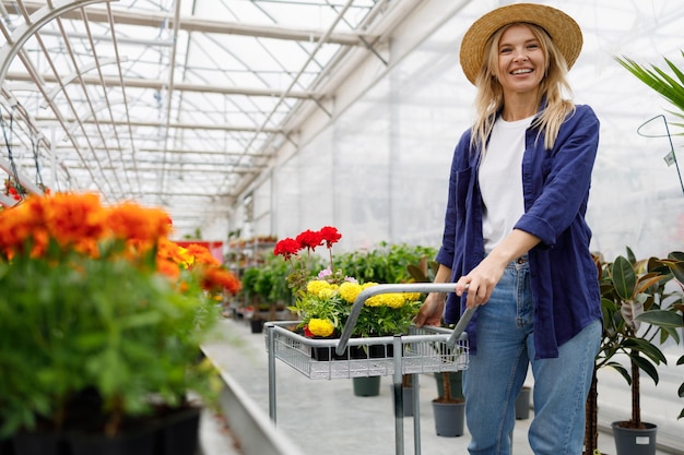 麦わら帽子をかぶったきれいな女性が、温室の周りに鉢植えの花でいっぱいのカートを押しています