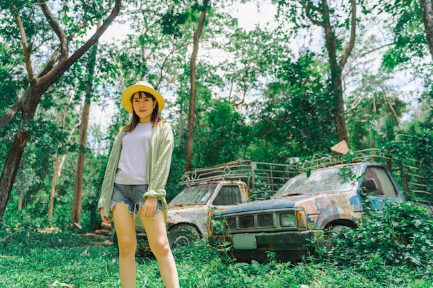 A pretty woman in a straw hat stood in the woods behind an old car parked