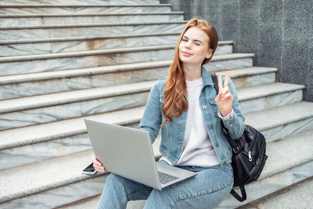 Pretty woman student with laptop sits on the stairs outdoors Distance education