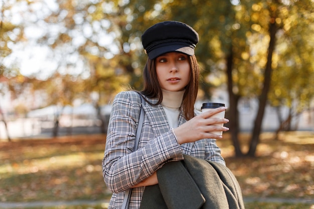 Красивая женщина в стильной кепке с модным костюмом держит в руках пальто и вкусный кофе и гуляет в осеннем парке