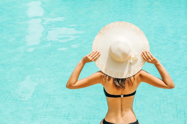 Pretty woman touching hat and enjoying a swimming pool at the resort