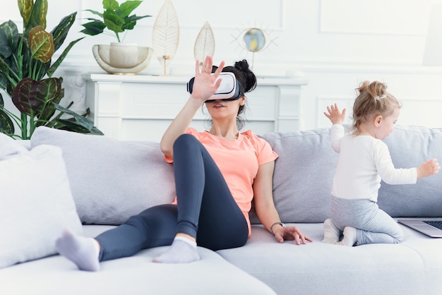 Pretty woman uses virtual reality goggles at home on sofa while her daughter watches cartoons on laptop