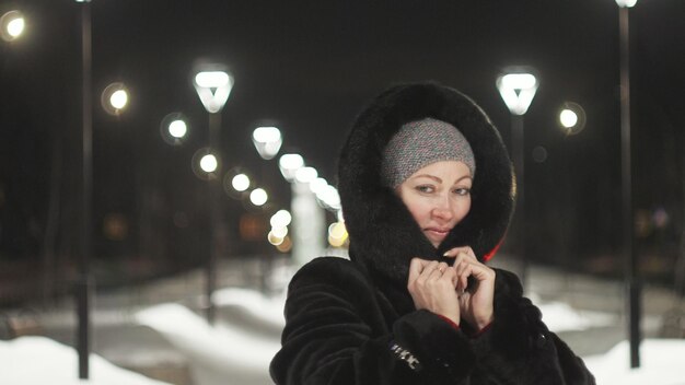 Pretty woman walking along the black fur coat outdoor girl\
walks on a winter night among the street lights of the city snow\
park with garlands beautiful sensual happy smile