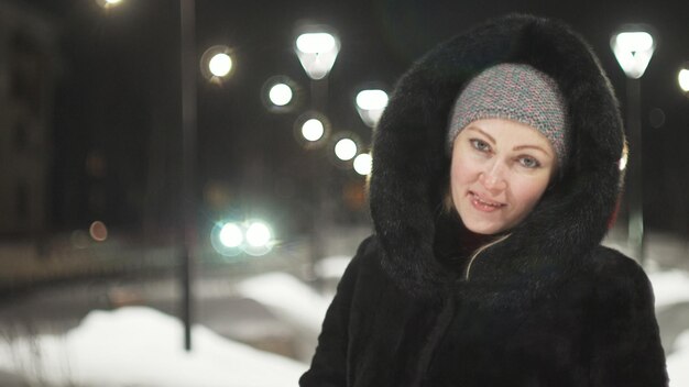 Pretty woman walking along the black fur coat Outdoor Girl walks on a winter night among the street lights of the city Snow park with garlands Beautiful sensual happy smile