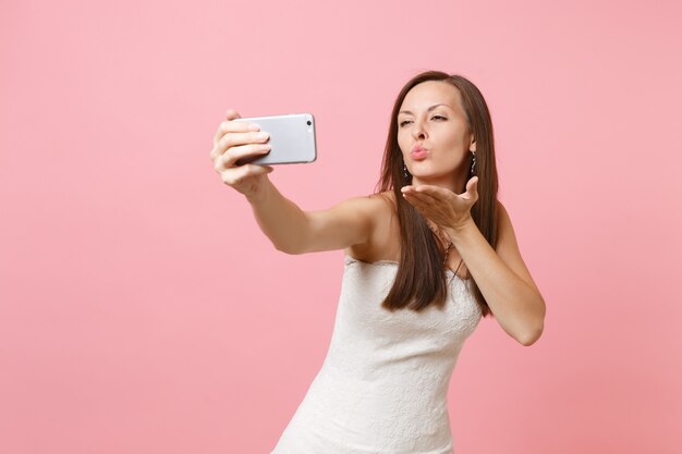 Pretty woman in white dress blowing lips, sending air kiss, doing taking selfie shot on mobile phone, making video call