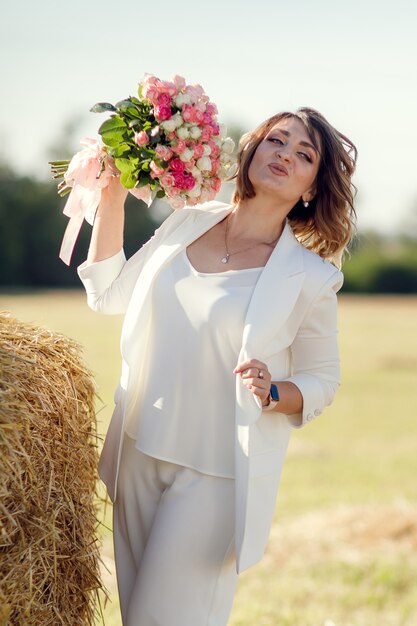 バラの花束を持つきれいな女性がフィールドの干し草の俵の近くに立っています。