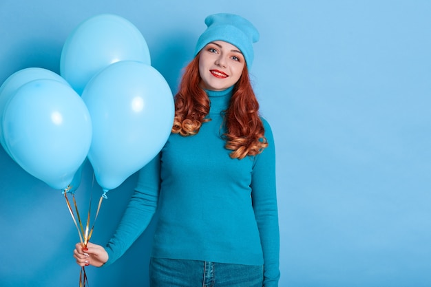 Pretty woman with bunch of balloons posing against blue wall with charming smile