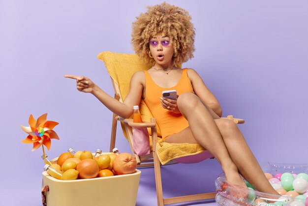 Pretty woman with curly haired woman wears sunglasses swimsuit\
holds mobile phone indicates away into distance shows something\
amazing keeps legs in inflated pool uses portable fridge full of\
fruits