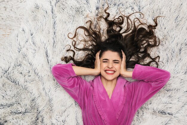 Pretty woman with long curly hair wearing girlish housecoat lying on white fur in apartment