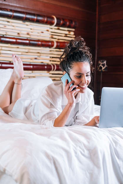 Pretty woman with phone and laptop in bed