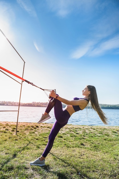 Красивая женщина в спортивной одежде делает отжимания в кроссфите с ремнями для фитнеса trx на открытом воздухе возле озера в дневное время. Здоровый образ жизни