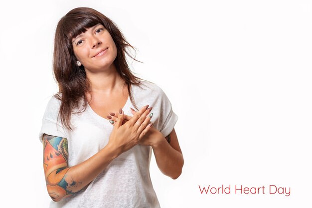 A pretty woman with tattoos on her arm holds her heart with her hands and smiles. White background. Copy space. The concept of world heart day.