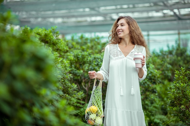 プリティ・ウーマン。野菜の袋と手にカップと白いドレスを着た女性