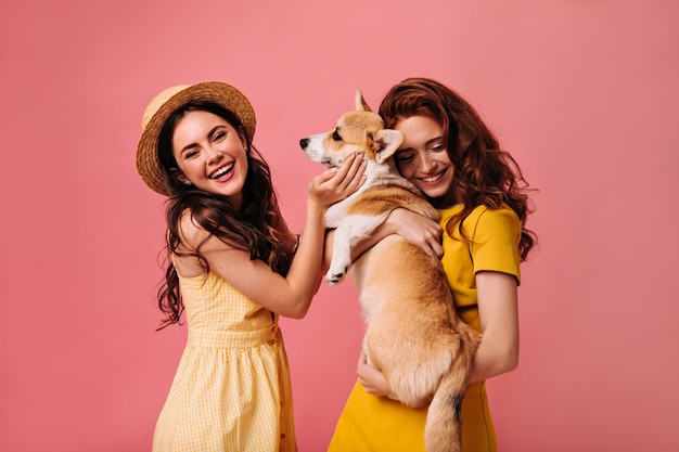 Pretty women hugging corgi on pink background Cheerful girls with curly hair and yellow dresses hold small funny dog and smile cute