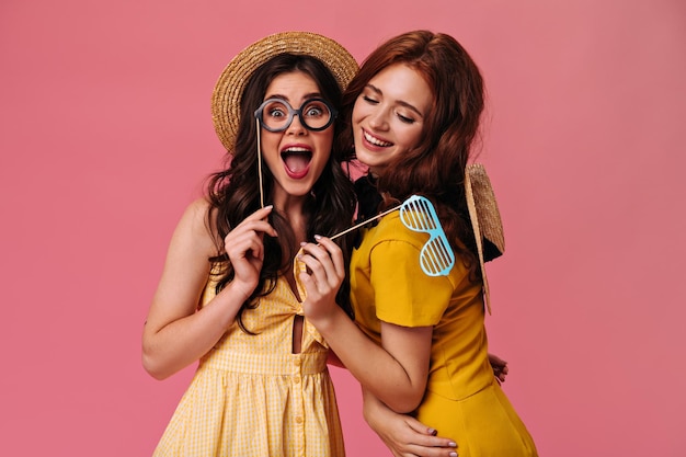 Pretty women laugh and hold eyeglasses on isolated background Wonderful girls in yellow summer dresses and hats show bright emotions
