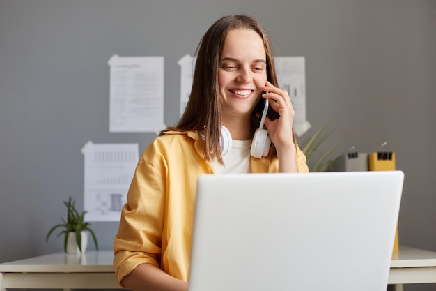 Pretty young adult woman working on laptop at her workplace in office talking with client with charming smile being pleased and friendly trying to sell her service or works in call center