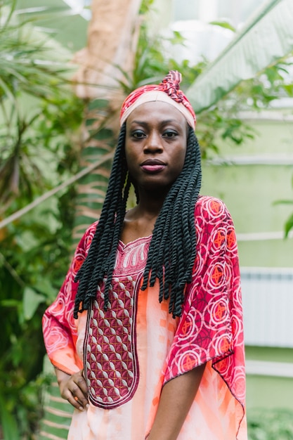 A pretty young Afro-American woman stands under palm trees in the jungle and smiles broadly in a national African costume.