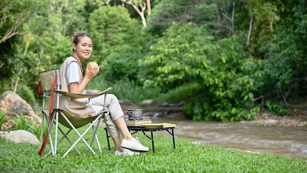 꽤 젊은 아시아 여성 캠퍼가 커피를 홀짝이며 숲 속의 강 근처에 앉아 있다