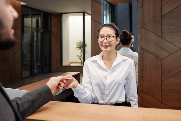 Pretty young Asian female receptionist passing card from hotel room to business traveler over reception counter