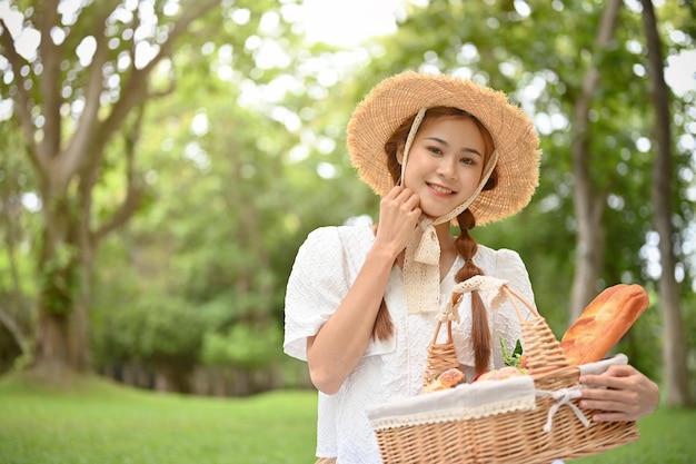 Femmina asiatica abbastanza giovane che indossa un cappello di paglia e che tiene un cestino di vimini dei pasticcini nel giardino