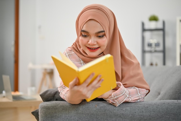 かなり若いアジアのイスラム教徒の女性が本を読んで、リビング ルームのソファに横になっています。