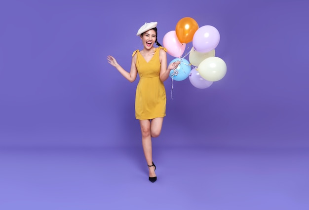 Pretty young asian woman at celebration party holding colourful balloon running and smile face. Happy new year or Birthday eve celebrating concept on bright purple wall.