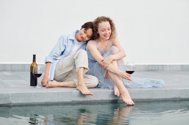 Pretty young Asian woman leaning on shoulder of her girlfriend when they are drinking red wine outdoors