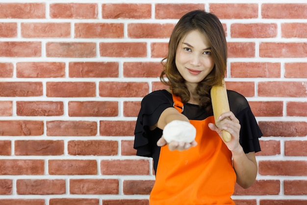 Pretty young Asian woman in orange apron smilingly stand in front of brick wall of kitchen and raising rolling pin and white dough to show that ready to begin funny cooking of kneading on holiday