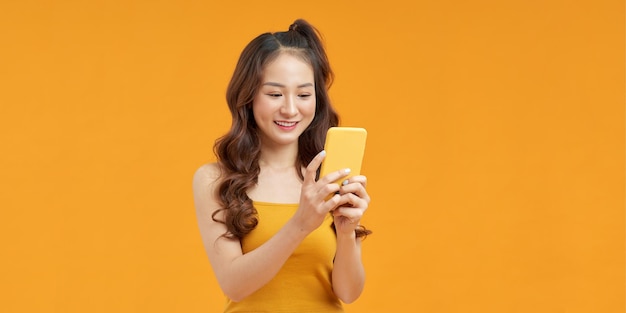 Pretty young asian woman using smartphone standing on isolated vivid yellow background