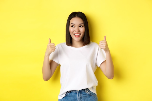 Довольно молодая азиатская женщина в белой футболке, показывая большие пальцы руки вверх и улыбаясь, хвалит хорошее предложение, рекомендует продукт, довольна стоя на желтом фоне.