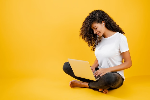 Pretty young black african woman sitting with laptop and earpods isolated over yellow 