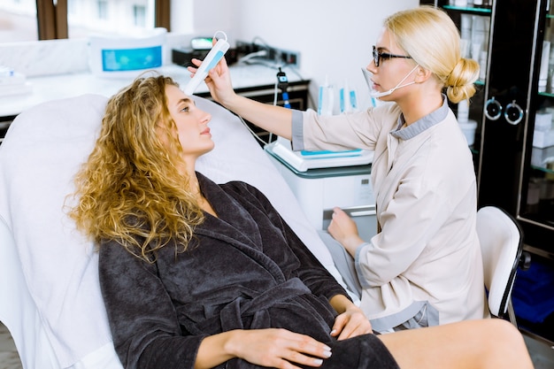 Pretty young blond curly woman during face skin diagnostic in the professional beauty salon with female doctor cosmetologist