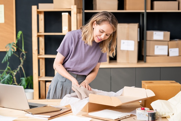 Pretty young blond female manager of online shop storage room bending over table and putting folded knitted sweater into cardboard box