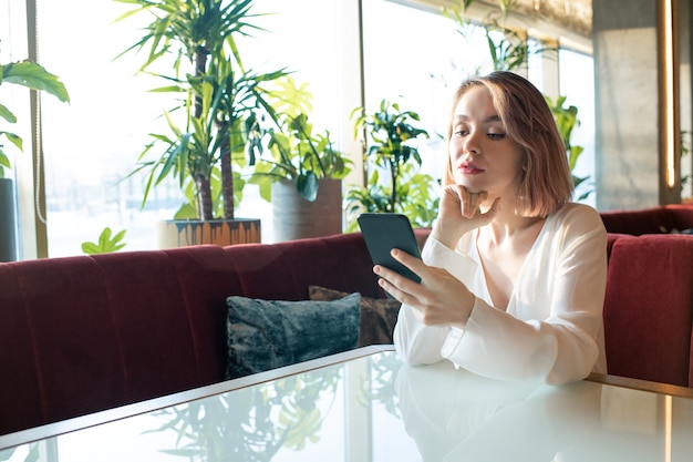 Pretty young blond woman in white silk blouse scrolling in smartphone while sitting by table on couch in comfortable restaurant