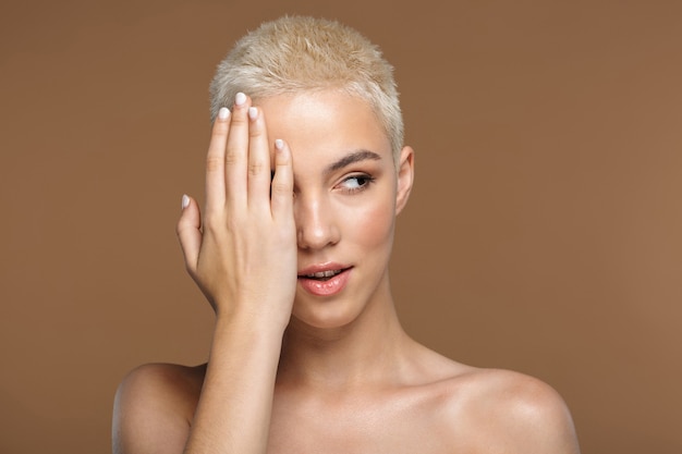 Una bella giovane donna bionda elegante con taglio di capelli corto in posa isolata sul muro beige scuro che copre metà del viso.