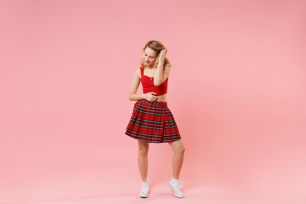 Pretty young blonde woman girl in red sexy clothes isolated on pastel pink background studio portrait. People sincere emotions lifestyle concept. Mock up copy space. Looking down, put hand on hair.