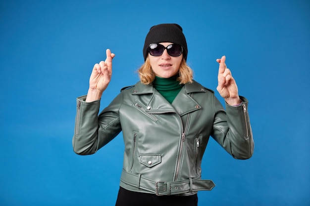 Pretty young blonde woman in green jacket posing isolated on blue background People lifestyle concept Mock up copy space Waiting for special moment keeping fingers crossed making wish