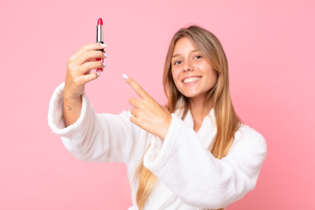 Pretty young blonde woman wearing bath robe and holding a lipstick