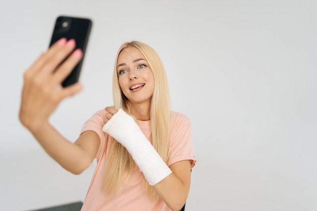 Pretty young blonde woman with broken arm wrapped in plaster bandage chatting mobile phone making video call taking selfie picture