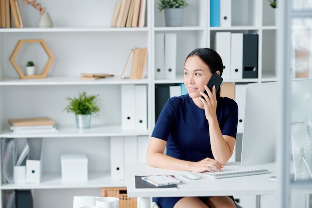 Pretty young brunette businesswoman with smartphone sitting by desk