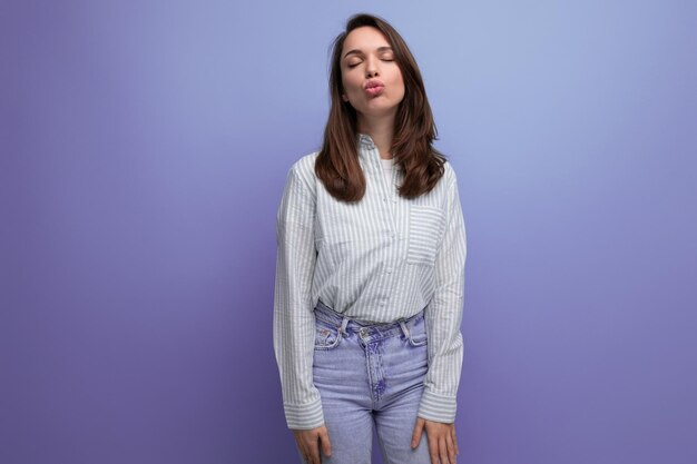 Pretty young brunette female adult in striped shirt and jeans sends air kiss isolated on plain