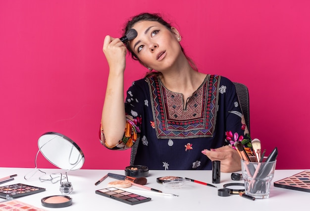 Pretty young brunette girl sitting at table with makeup tools applying blush with makeup brush looking up isolated on pink wall with copy space